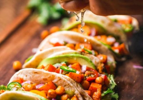 A hand squeezes lime over tacos filled with chickpeas, vegetables, and cilantro. Lime slices and cherry tomatoes are on the wooden board.