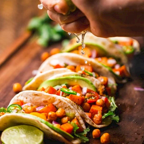 A hand squeezes lime over tacos filled with chickpeas, vegetables, and cilantro. Lime slices and cherry tomatoes are on the wooden board.