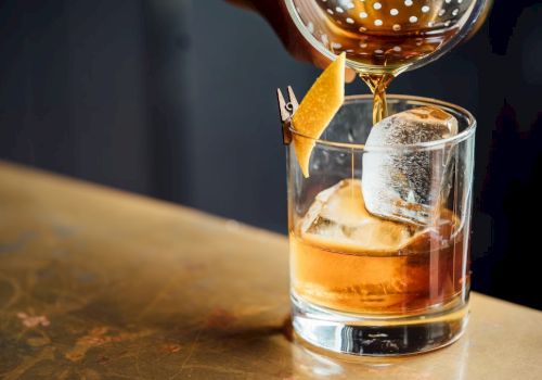A drink is being poured into a glass with large ice cubes, garnished with a citrus peel for a stylish presentation.