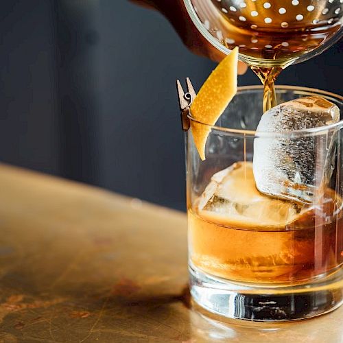A drink is being poured into a glass with large ice cubes, garnished with a citrus peel for a stylish presentation.