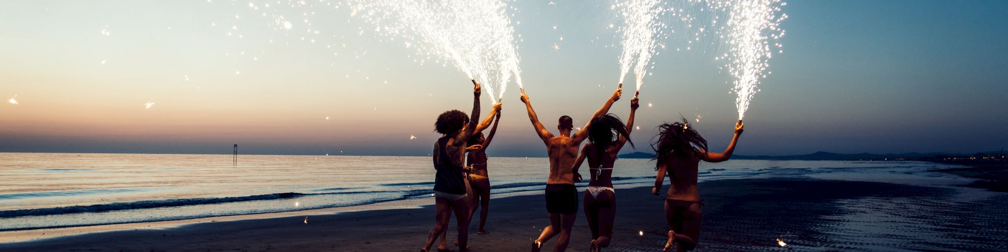 A group of people on a beach at dusk, holding sparklers that light up the scene with bright trails of sparks.