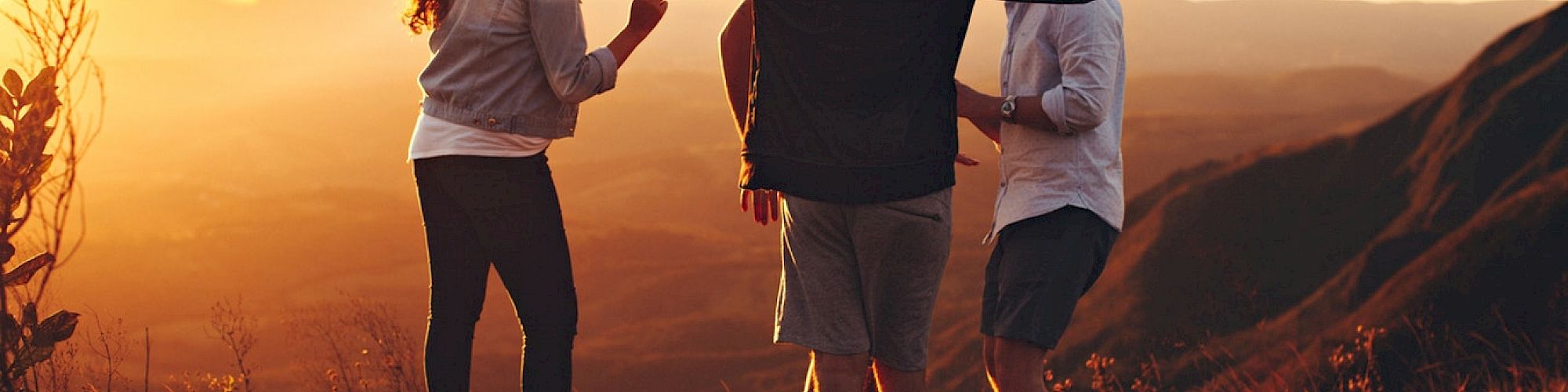 A group of people celebrating at sunset on a grassy hill, with mountains in the background, creating a vibrant and joyful atmosphere.