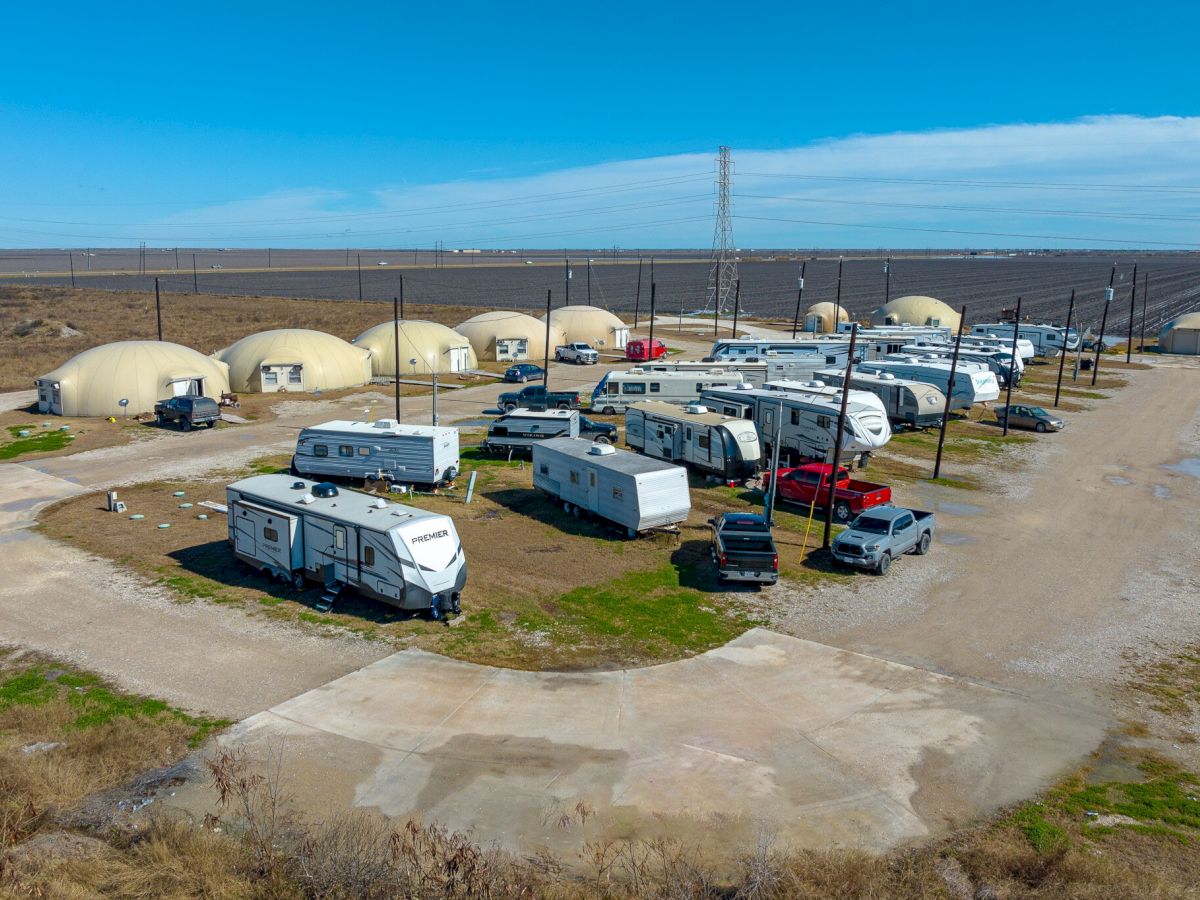 The image shows a parking area with numerous RVs and trailers, alongside dome-shaped structures in an open field landscape.