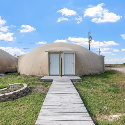 Two dome-shaped buildings with double doors are connected by wooden walkways, set against a clear blue sky.