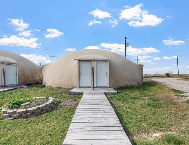 Two dome-shaped buildings with double doors are connected by wooden walkways, set against a clear blue sky.