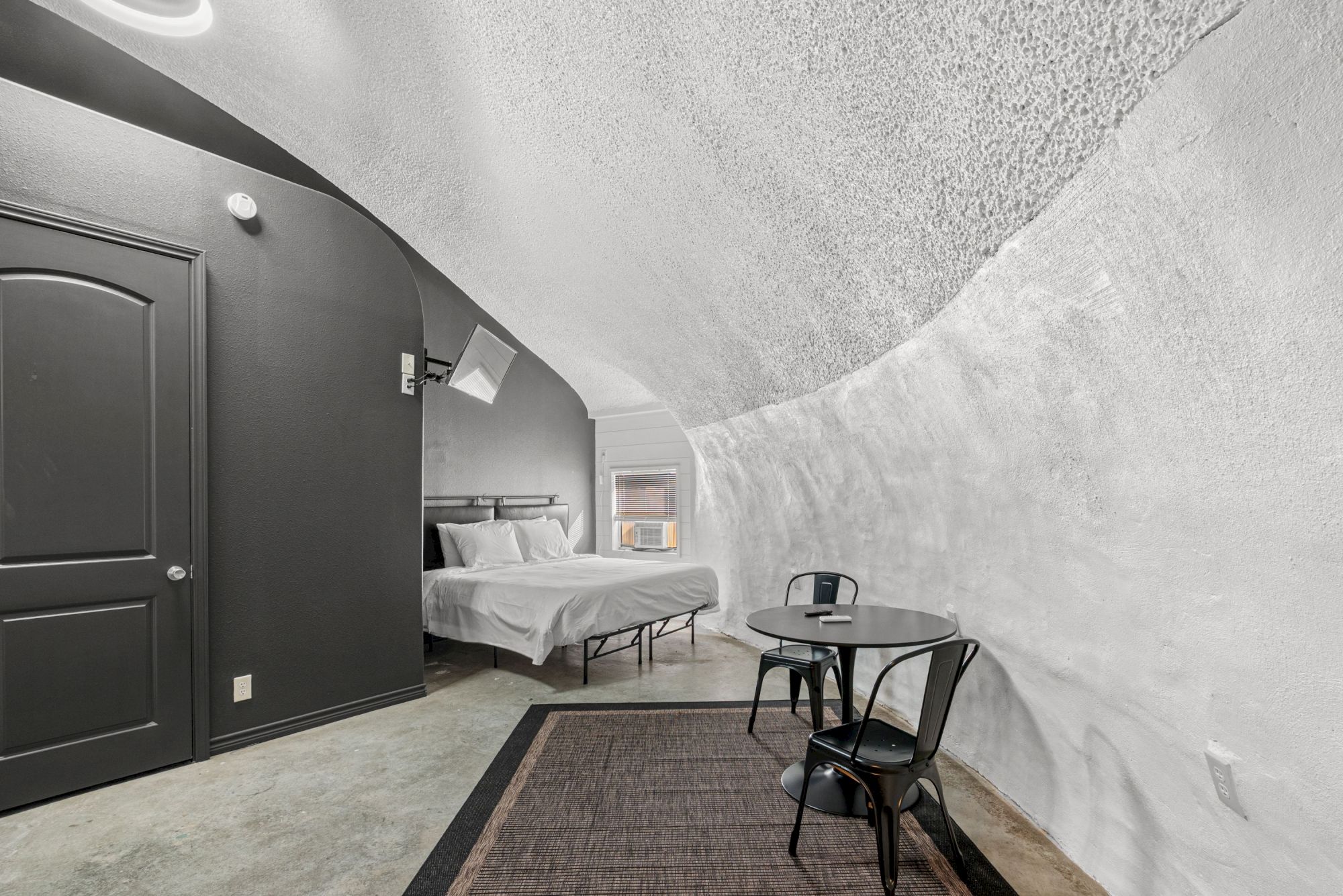 A minimalist room with a curved white ceiling, bed, small table with two chairs, gray door, and textured walls, creating a cozy atmosphere.