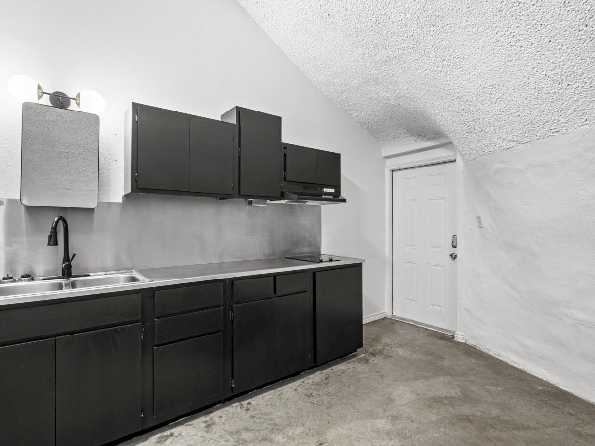 A minimalist kitchen with black cabinets and stainless steel sink, set against white walls and ceiling, with a closed door in view.