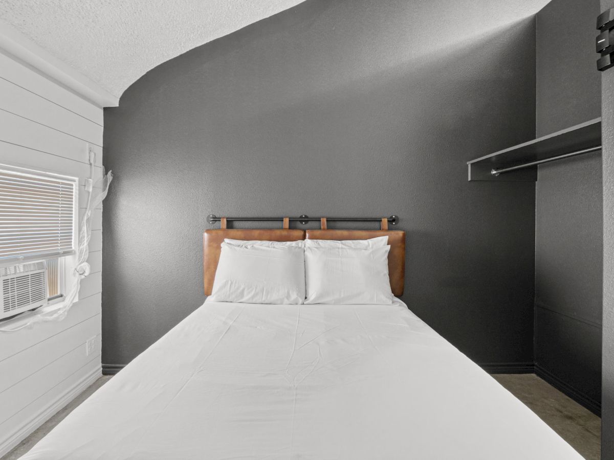 The image shows a minimalist bedroom with a bed, white bedding, gray walls, and a window with blinds on the side.