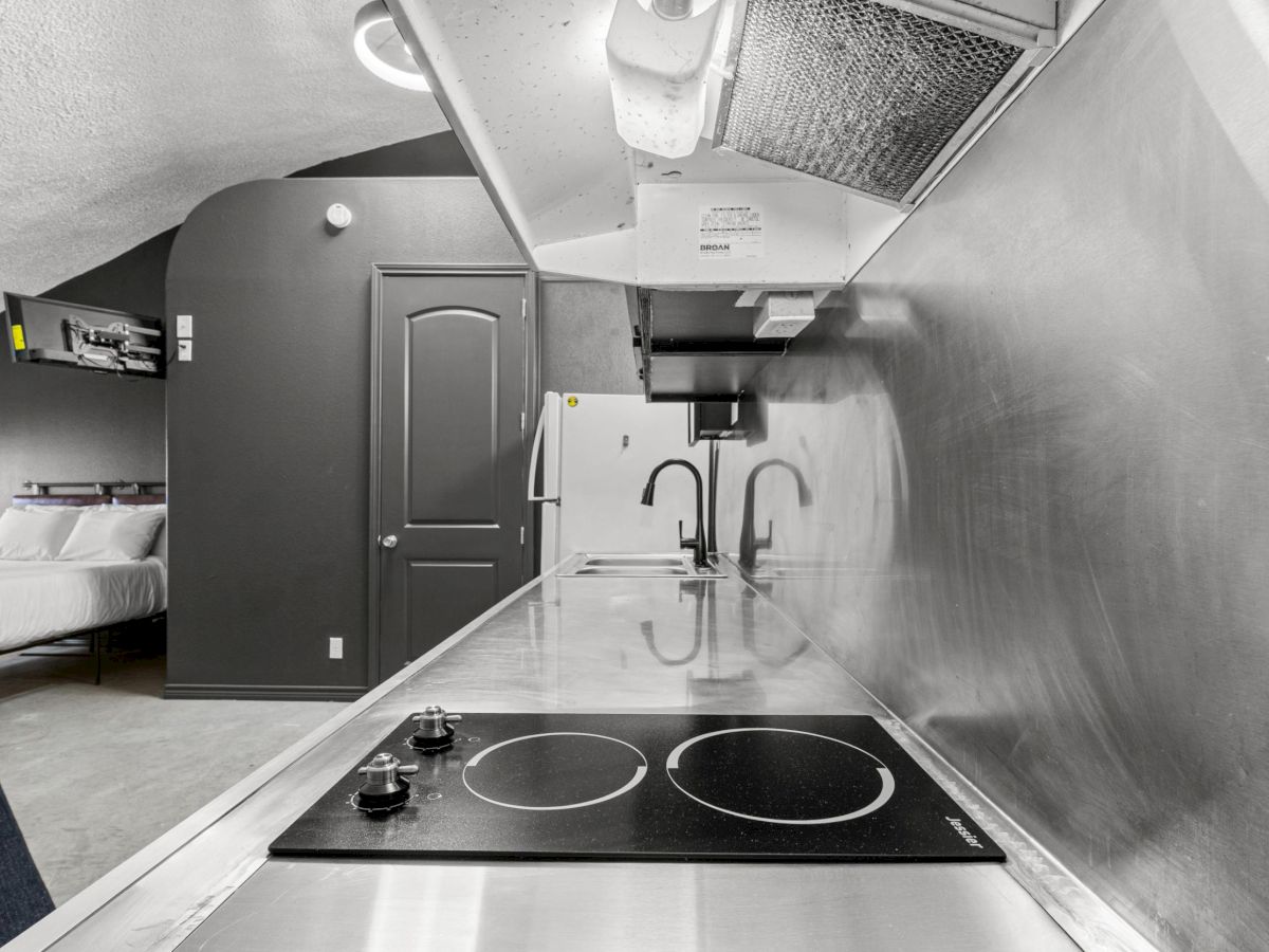 The image shows a modern kitchen area with a stovetop, metallic surfaces, and a view into an adjoining room with a bed.