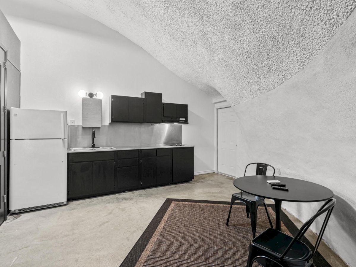 A minimalist kitchen in a cave-like room with black cabinets, a small dining table, and chairs; fridge and lights are visible.