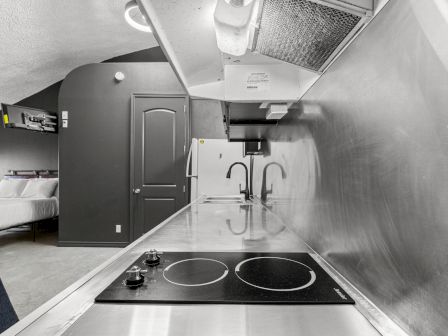 A sleek stainless steel kitchen with a two-burner stove, modern sinks, and a view into a bedroom area.