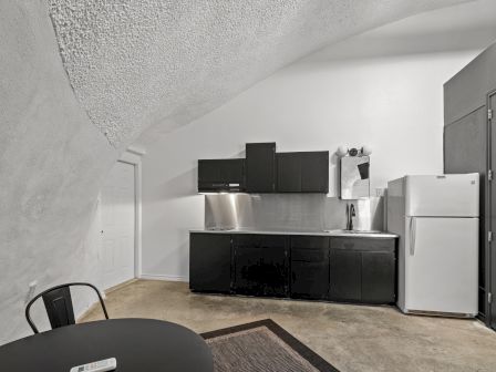 A minimalist kitchen with black cabinetry, a refrigerator, and a small dining area under a curved ceiling.